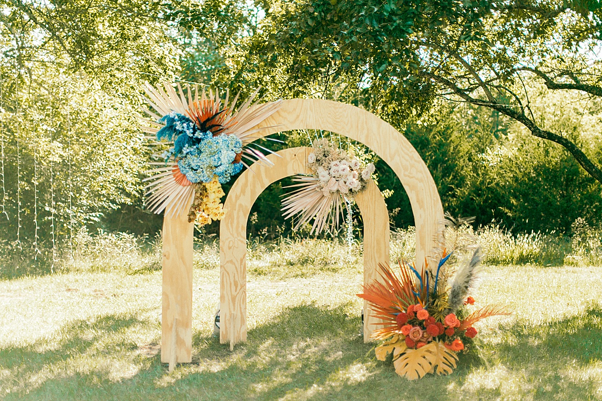 Double wooden arch wedding ceremony backdrop with large blue and orange boho floral sprays.